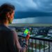 Person on an Airdrie balcony at dusk looking toward storm clouds with a smartphone casting subtle radar-like colors, suggesting real-time weather awareness.