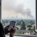 Person holding a smartphone near an open window as wildfire smoke fills the neighborhood air.