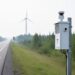An air-quality monitoring station near a roadside with wind turbines in the distance under overcast daylight, suggesting local air quality tracking.